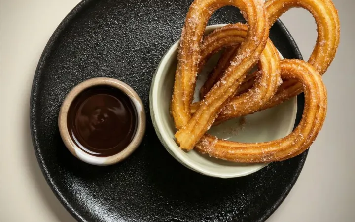 Churros cu sos de ciocolată pe farfurie neagră, pe fundal gri deschis