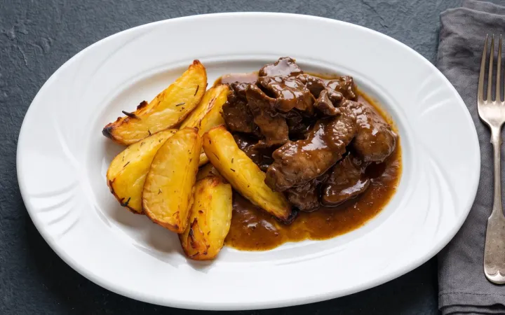 Carne de vită în sos cu cartofi wedges pe o farfurie ovală albă, pe fundal închis de ardezie, cu furculiță și șervet gri