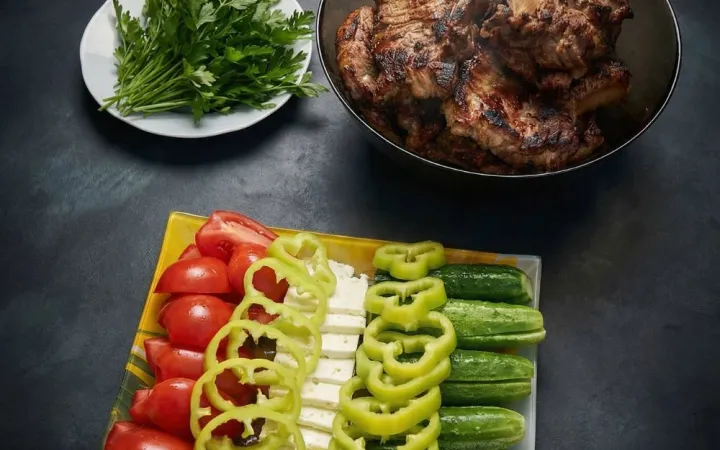 Platou cu legume feliate, verdeață și un bol cu carne la grătar, pe fundal închis de ardezie